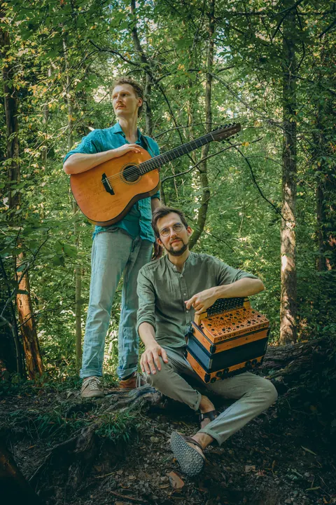 Press photo: Timmy standing with guitar, Jonny sitting on a log with the accordion